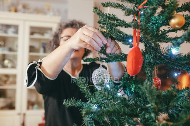 Акцентированная женщина средних лет в очках с наклонной головой, украшающей елку перед новогодним праздником в доме — стоковое фото