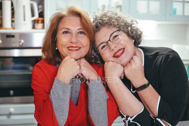 Heureuse femme lesbienne d'âge moyen appuyée sur l'épaule d'une femme souriante bien-aimée dans la maison regardant la caméra — Photo de stock