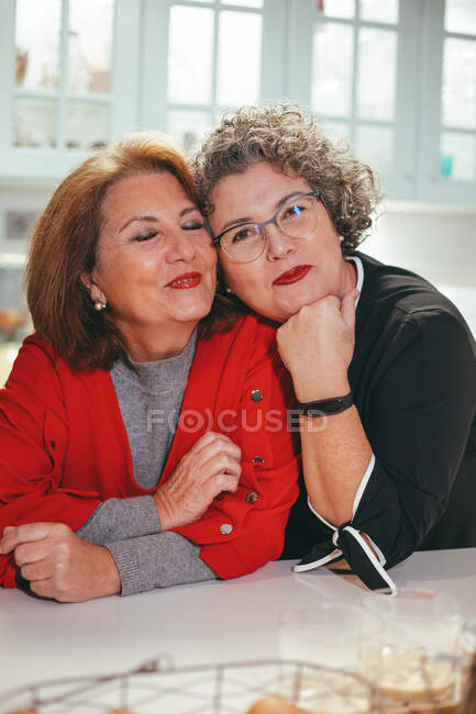 Frohe lesbische Frau mittleren Alters lehnt auf der Schulter einer lächelnden Geliebten im Haus und schaut in die Kamera — Stockfoto