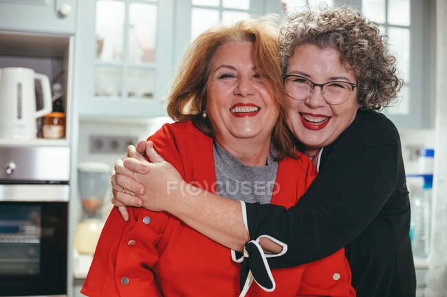 Glad middle aged lesbian woman embracing smiling female beloved in house looking at camera — Stock Photo