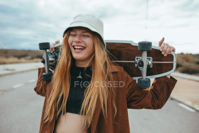 Allegro giovane donna millenaria in abito elegante e cappello ridere ad occhi chiusi mentre in piedi su strada asfaltata con skateboard dietro la testa dopo la guida — Foto stock