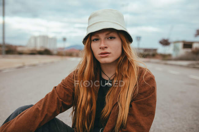 Jeune femme confiante avec de longs cheveux roux assis dans la rue par temps nuageux et regardant la caméra — Photo de stock