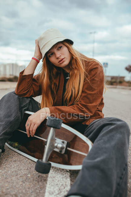Joven patinadora femenina con el pelo largo y rubio en traje de moda sentada en la carretera de asfalto con el patín cruiser en la mano contra el cielo nublado en el campo - foto de stock