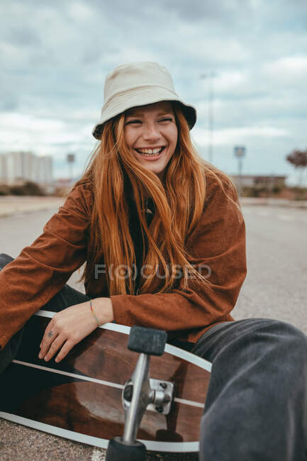 Cheerful young female millennial in stylish outfit and hat laughing with closed eyes while sitting on asphalt road with skateboard behind head after riding — Stock Photo