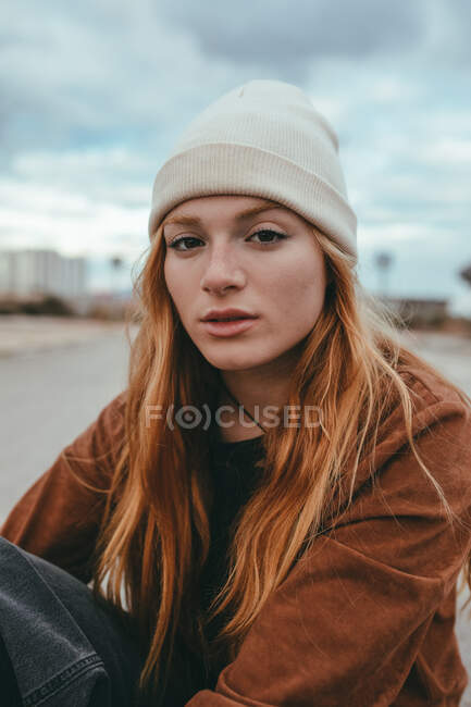 Jeune femme confiante avec de longs cheveux roux debout sur la rue par temps nuageux et regardant la caméra — Photo de stock
