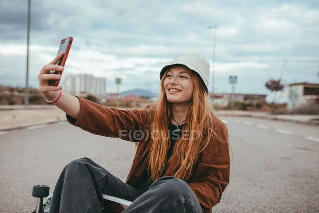 Glückliche Teenagerin mit langen roten Haaren im trendigen Outfit und Hut sitzt auf dem Skateboard und lächelt beim Selfie mit dem Handy — Stockfoto