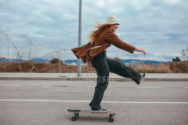 Vista lateral de la joven feliz millennial en traje elegante y sombrero haciendo truco en el monopatín mientras se monta en el camino de asfalto contra el cielo nublado en el campo - foto de stock
