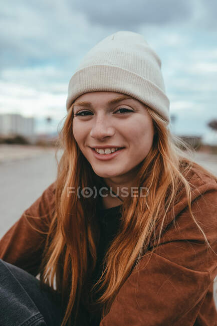 Cheerful young female with long ginger hair sitting on street on cloudy day and looking at camera — Stock Photo