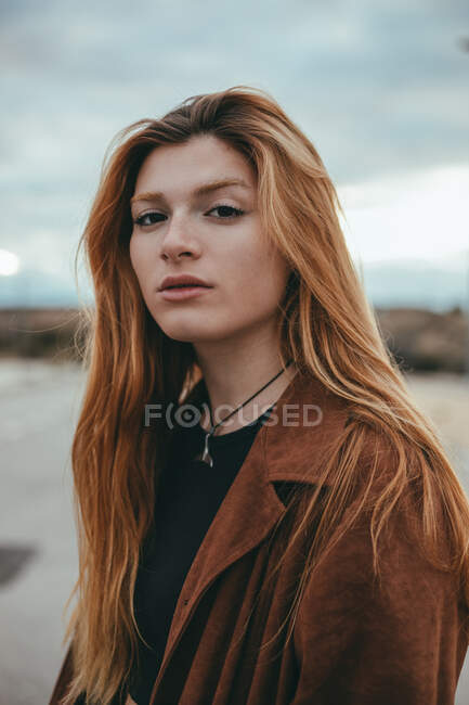 Mulher jovem confiante com cabelos longos de gengibre em pé na rua no dia nublado e olhando para a câmera — Fotografia de Stock