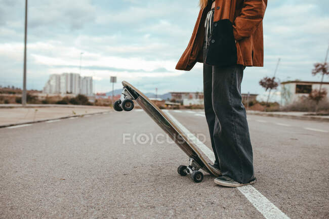Side view of crop female skater in trendy outfit standing on asphalt road with cruiser skateboard in countryside — Stock Photo