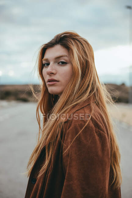 Vista lateral de mujer joven y segura con el pelo largo de jengibre de pie en la calle en el día nublado y mirando a la cámara - foto de stock
