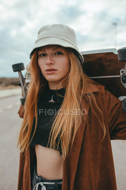 Young female skater with long blond hair in trendy outfit standing on asphalt road with cruiser skateboard in hand against cloudy sky in countryside — Stock Photo