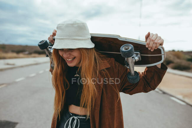 Весела молода жінка тисячоліття в стильному вбранні і капелюсі сміється з закритими очима, стоячи на асфальтній дорозі зі скейтбордом за головою після їзди — стокове фото