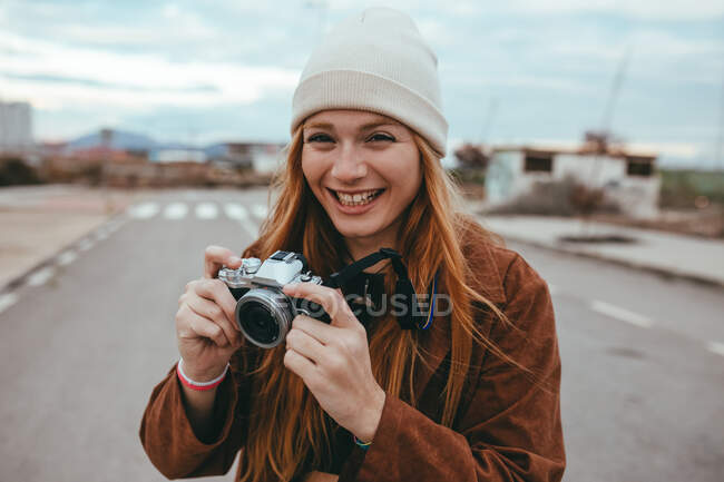 Giovane donna positiva con lunghi capelli rossicci in abiti eleganti e cappello sorridente mentre in piedi sulla strada con fotocamera vintage in mano durante il viaggio in campagna — Foto stock