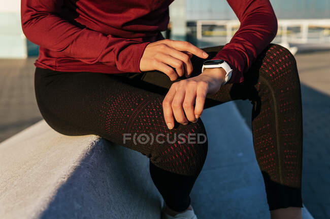 Schnuppern anonyme Sportlerin in stylischer Sportbekleidung sitzt am Rand und überprüft den Puls auf Fitness-Tracker, während sie während des Outdoor-Trainings in der Stadt ruht — Stockfoto