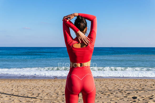 Back view female athlete in red sportswear standing on sandy beach near wavy ocean and stretching arms before training — Stock Photo