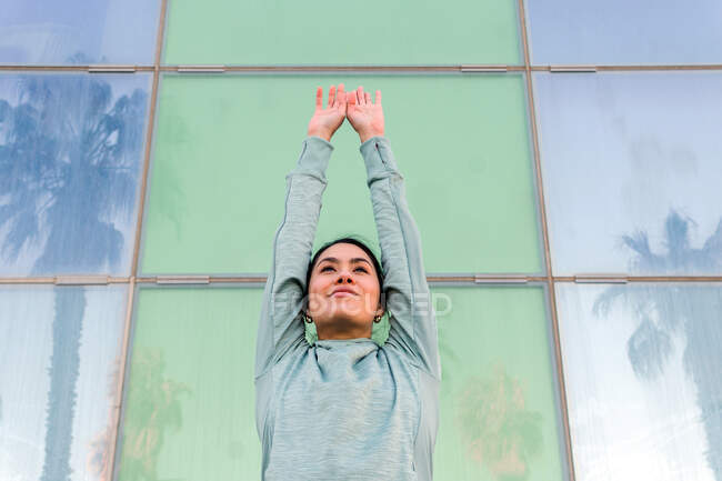 Уверенная в себе молодая спортсменка в стильной спортивной одежде смотрит в сторону современного стеклянного здания на городской улице — стоковое фото