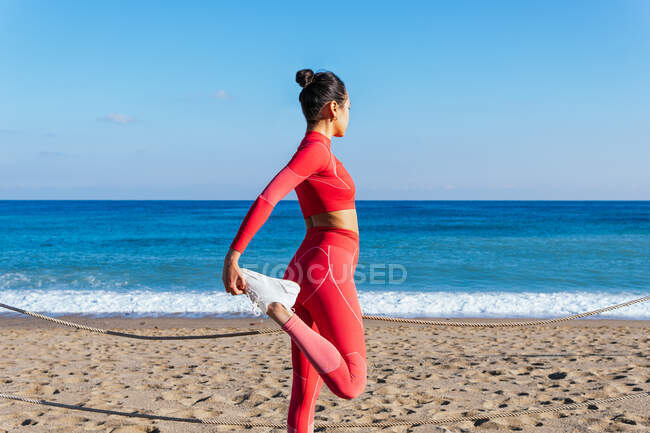 Вид збоку на невпізнавану молоду етнічну леді в спортивному одязі та кросівках, що розтягують ноги, стоячи на піщаному морському пляжі під час тренування на відкритому повітрі — стокове фото
