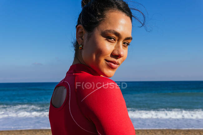 Vue latérale de la jeune sportive hispanique aux cheveux foncés en vêtements de sport rouges souriant et regardant la caméra pendant l'entraînement sur une plage de sable contre un ciel bleu sans nuages — Photo de stock