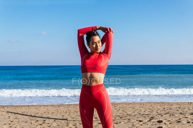 Самоуверенная молодая спортсменка в красной спортивной одежде стоит на песчаном пляже возле волнистого океана и вытягивает руки перед тренировкой — стоковое фото