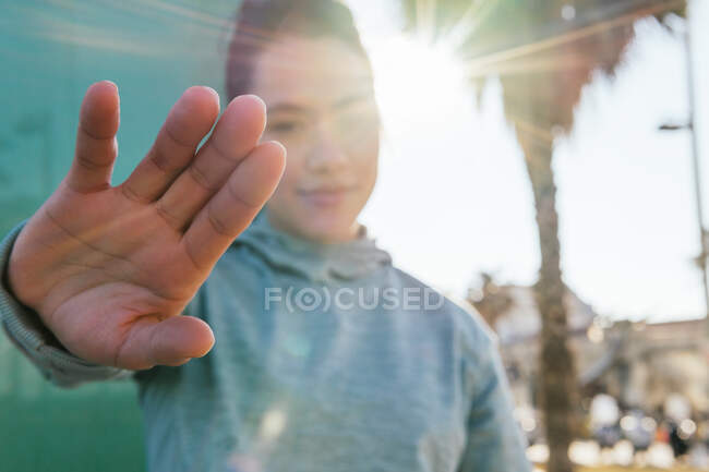 Young ethnic female in casual clothes covering face from bright sunlight with hand and looking at camera while standing near palms on city street — Stock Photo
