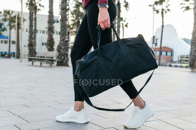 Вид збоку врожаю підходить жінці в модному спортивному вбранні і кросівках, що несуть сумочку під час прогулянки на брукованій міській площі — стокове фото