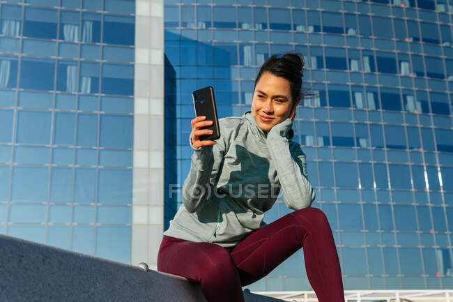 Glückliche junge hispanische Dame in Sportkleidung, die lächelt und ihr Handy benutzt, während sie sich nach dem Outdoor-Training an einem sonnigen Tag auf einer Bank in der Nähe eines modernen Gebäudes ausruht — Stockfoto