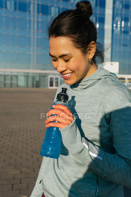 Calm young ethnic female athlete with dark hair in sportswear drinking water from bottle during outdoor workout at seaside on sunny day — Stock Photo