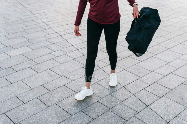 Crop fit female in trendy sportive outfit and sneakers carrying handbag while walking on paved city square - foto de stock