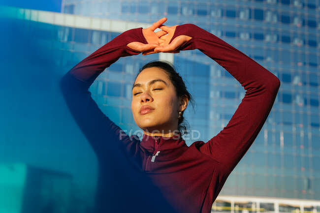Confident young ethnic female athlete in stylish sportswear with closed eyes standing against modern glass building on city street — Stock Photo
