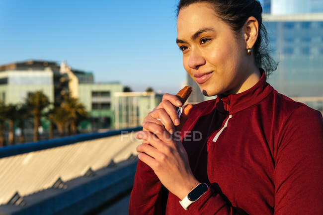 Positive young ethnic female athlete in sportswear and fitness tracker having break and eating protein bar after outdoor training in city — Stock Photo