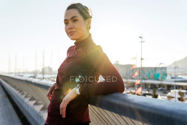 Positive self assured young ethnic female in trendy sportswear with fitness tracker leaning against fence and looking away while relaxing after outdoor training on urban embankment — Stock Photo