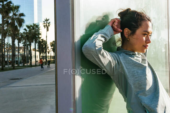 Впевнена молода етнічна жінка-спортсменка в стильному спортивному одязі, яка дивиться далеко, стоячи на тлі сучасного скляного будинку на міській вулиці — стокове фото