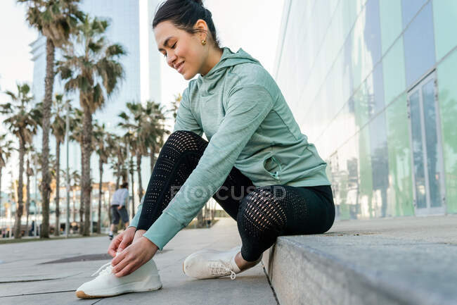 Vista lateral de la joven atleta étnica sonriente en ropa deportiva atando zapatillas durante el entrenamiento al aire libre en el terraplén de la ciudad en un día soleado - foto de stock