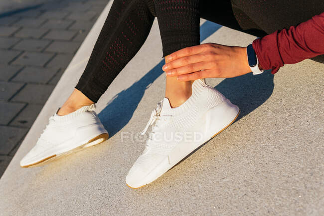 Вид збоку анонімної жінки в трендових білих кросівках і спортивному вбранні з трекером для фітнесу, що сидить на бетонній межі, маючи перерву під час тренувань на відкритому повітрі — стокове фото
