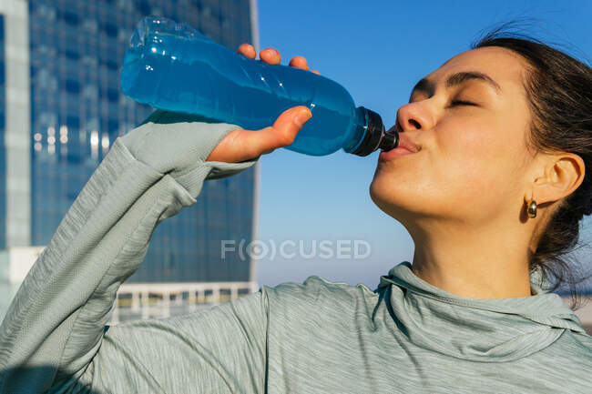 Спокійна молода етнічна жінка-спортсменка з темним волоссям у спортивному одязі п'є воду з пляшки під час тренування на відкритому повітрі на морі в сонячний день — стокове фото