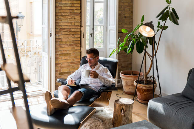 Empresário sério sentado em poltrona em casa e escrevendo planos no organizador enquanto desfruta de café durante o intervalo — Fotografia de Stock