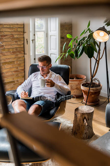 Серьезный бизнесмен сидит дома в кресле и пишет планы в органайзере, наслаждаясь кофе во время перерыва — стоковое фото