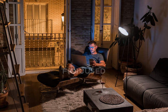 Busy male entrepreneur sitting in armchair and working on project while using laptop and smartphone — Stock Photo
