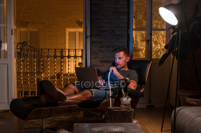 Homme détendu assis dans un fauteuil confortable avec ordinateur portable et fumer narguilé tout en regardant un film et profiter du week-end — Photo de stock