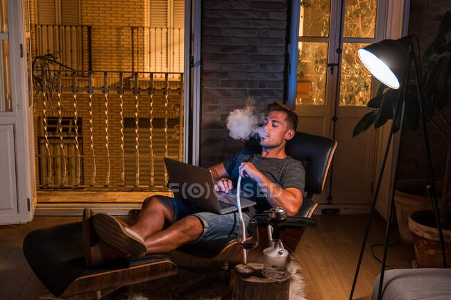 Rilassato maschio seduto in comoda poltrona con computer portatile e fumare narghilè durante la visione di film e godersi il fine settimana — Foto stock