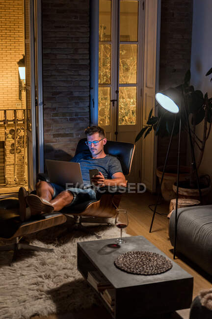 Entrepreneur masculin occupé assis dans un fauteuil et travaillant sur un projet tout en utilisant un ordinateur portable et un smartphone — Photo de stock