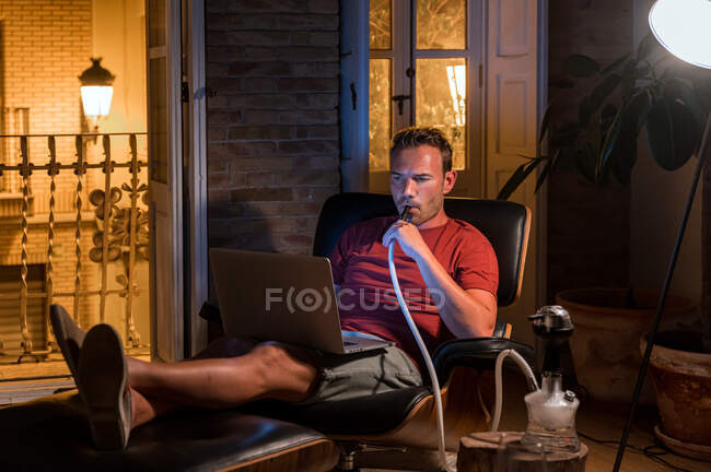 Relajado hombre sentado en un cómodo sillón con portátil y fumar narguile mientras ve la película y disfruta del fin de semana - foto de stock