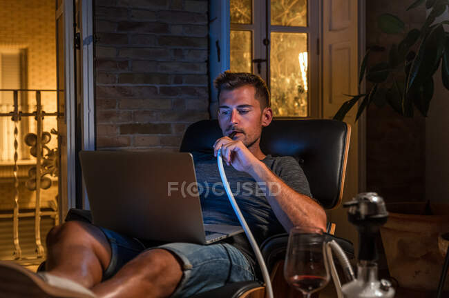 Relaxado macho sentado em poltrona confortável com laptop e fumar narguilé enquanto assiste filme e desfrutar de fim de semana — Fotografia de Stock