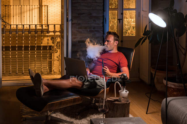 Розслаблений чоловік сидить у зручному кріслі з ноутбуком і курить кальян, дивлячись фільм і насолоджуючись вихідними — стокове фото