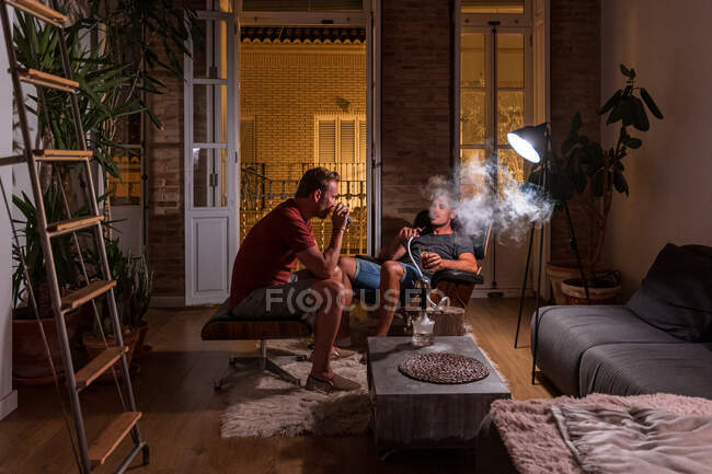 Amigos varones sentados en la sala de estar y descansando por la noche mientras beben alcohol y fuman narguile - foto de stock