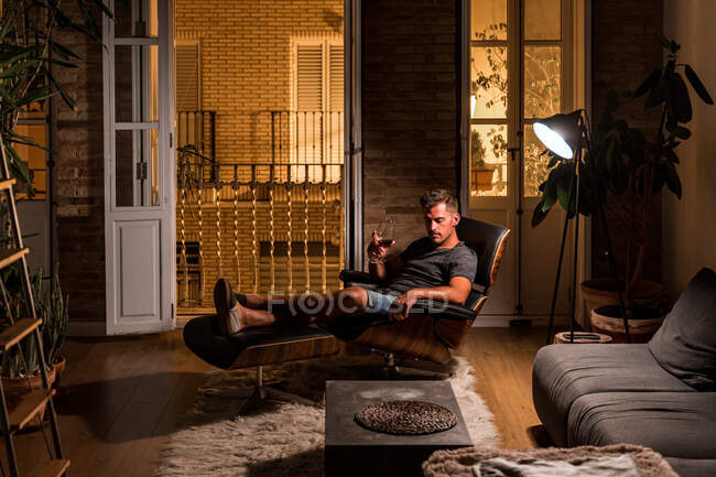 Вид сбоку мужчины в домашней одежде, сидящего в удобном кресле и пьющего красное вино, наслаждаясь вечером в темной комнате — стоковое фото