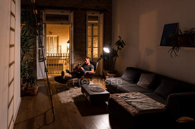 Seitenansicht eines Mannes im häuslichen Outfit, der im bequemen Sessel chillt und Rotwein trinkt, während er den Abend im dunklen Raum genießt — Stockfoto