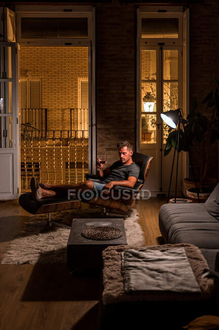 Вид сбоку мужчины в домашней одежде, сидящего в удобном кресле и пьющего красное вино, наслаждаясь вечером в темной комнате — стоковое фото