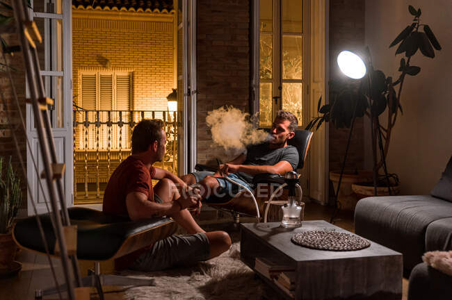 Hombres relajados sentados en la acogedora sala de estar y fumar narguile juntos mientras disfrutan de la noche en fin de semana - foto de stock